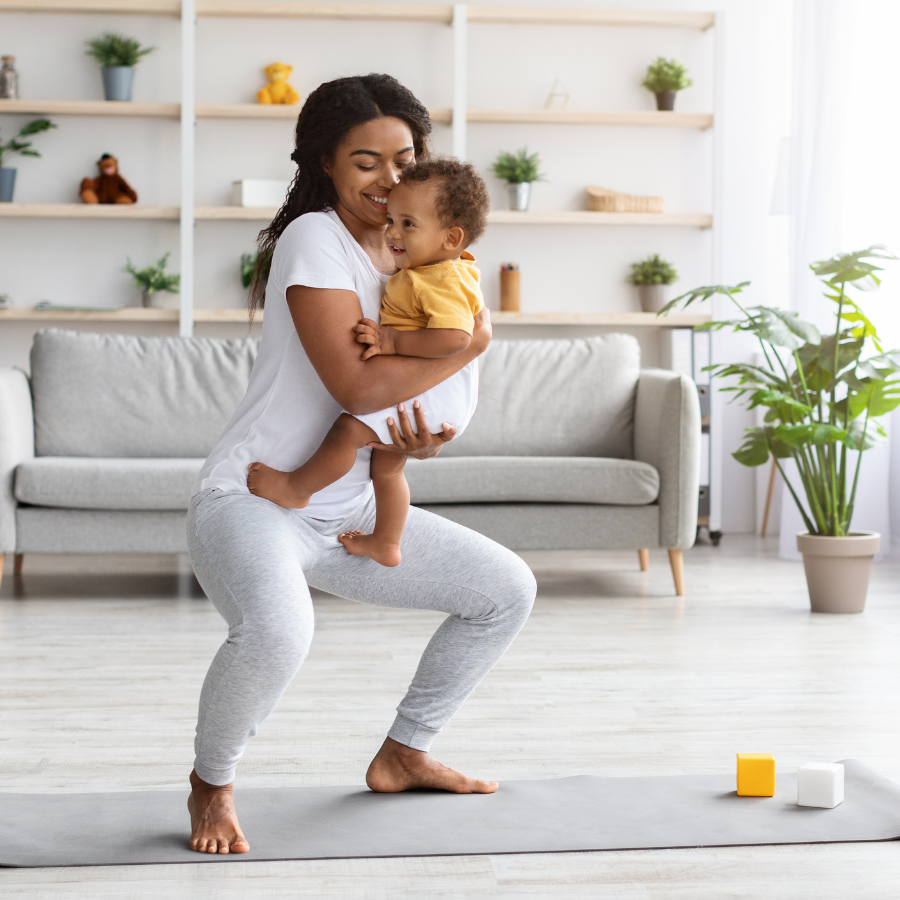 Mother doing postpartum exercise at home while holding her baby, practicing squats on a yoga mat in a bright living room.