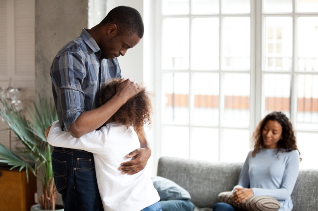 A father gently hugs his child while the co-parent watches calmly in the background, portraying the emotional care and cooperation needed for successful co-parenting transitions.